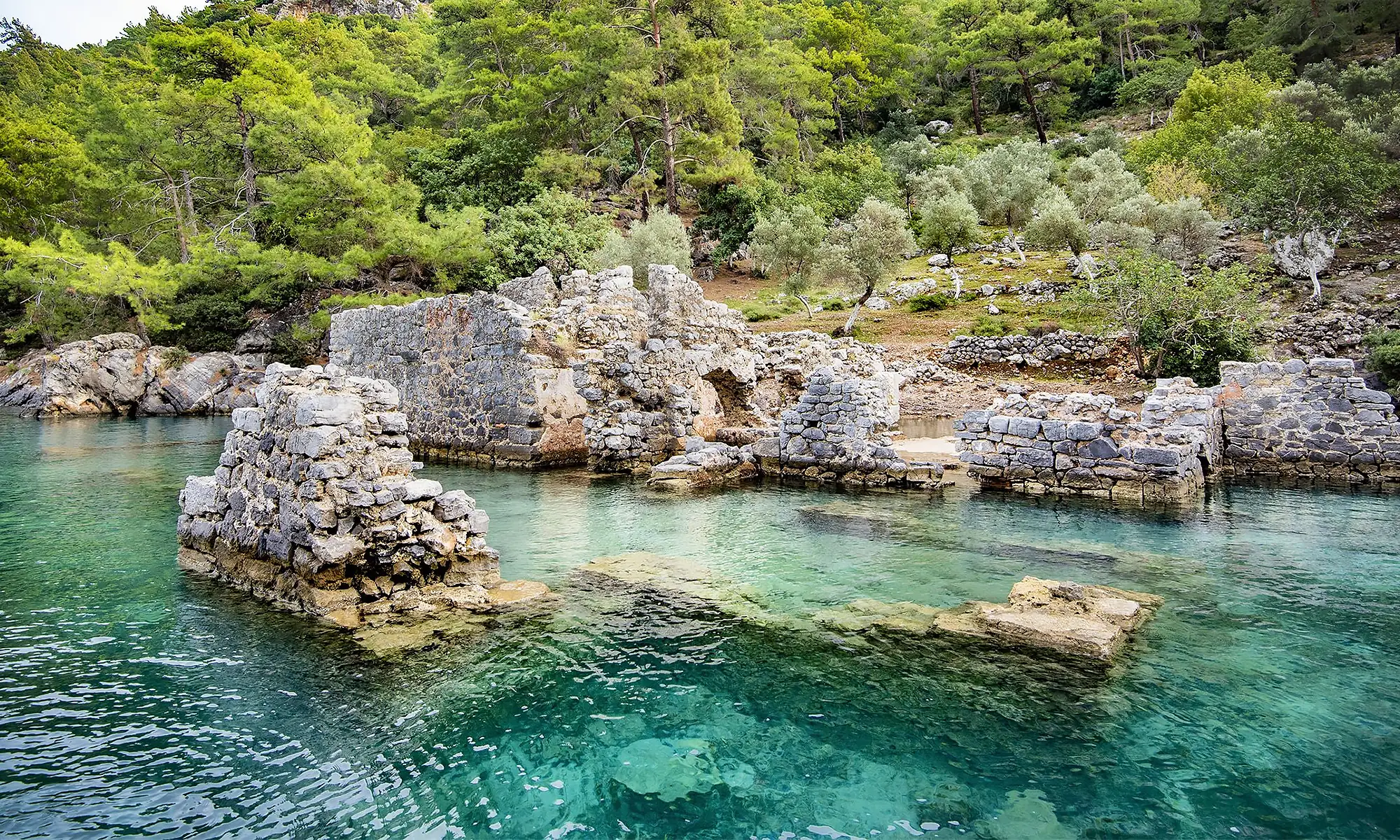 Göcek'teki Kleopatra Hamam Koyu'nun turkuaz suları ve batık manastır kalıntıları. Mavi yolculuk tekneleri için popüler yüzme ve tarih noktası.