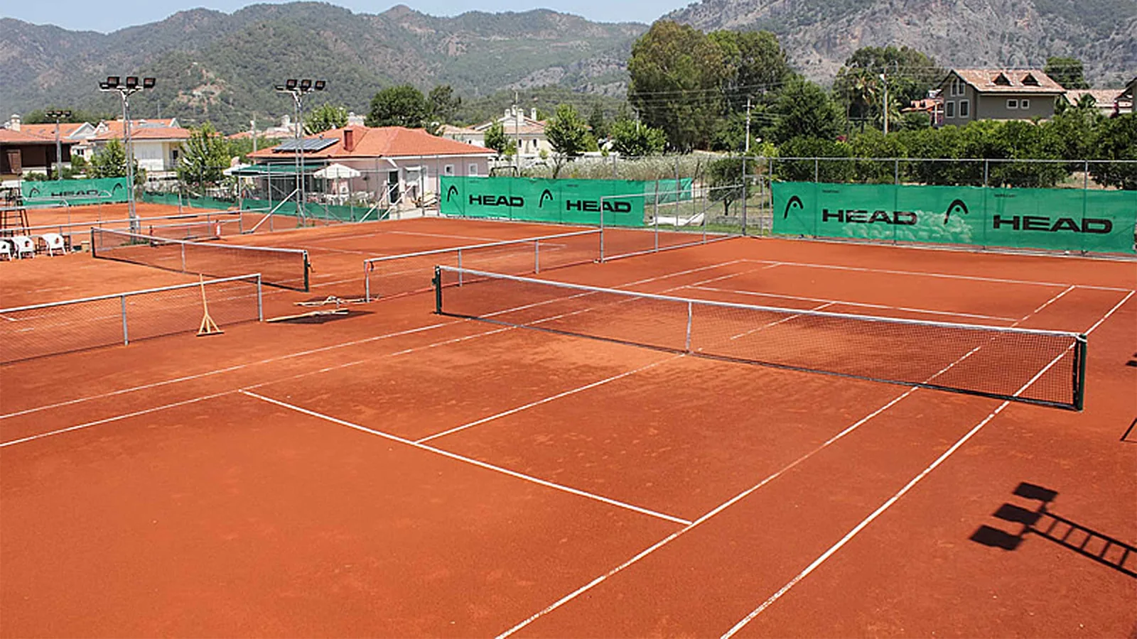 Göcek'teki bakımlı toprak tenis kortları ve arkasındaki dağ manzarası. Tatilciler ve yerel halk için spor ve rekreasyon imkanı sunan tesis.