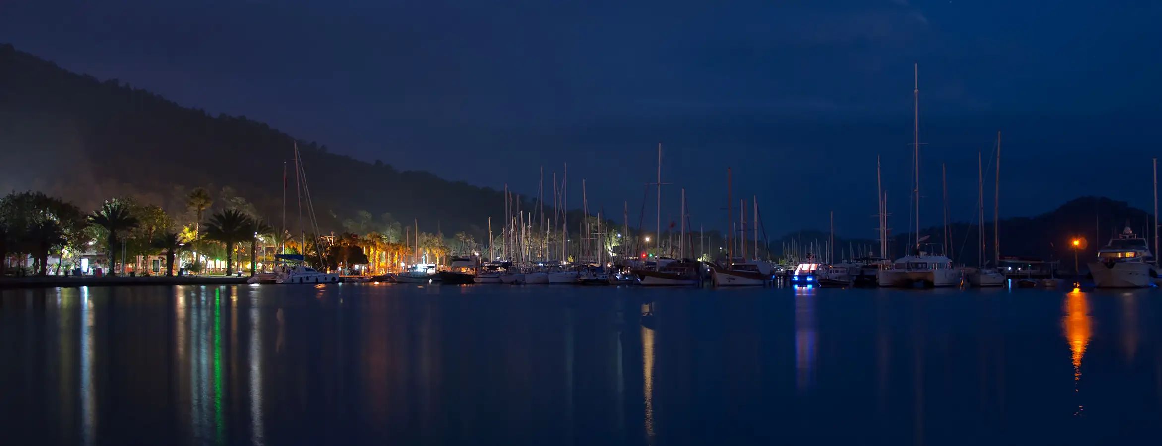 Göcek'in akşam saatlerindeki panoramik görünümü; marinaların ve kasabanın ışıkları, denize yansımaları. Gün batımında teknelerin ve sahilin büyüleyici manzarası.