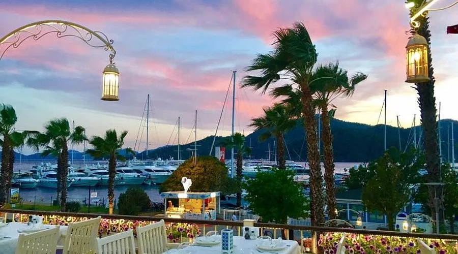 Göcek'te bir restoranın terasından gün batımı manzarası; denize yansıyan renkler ve teknelerin silüetleri.