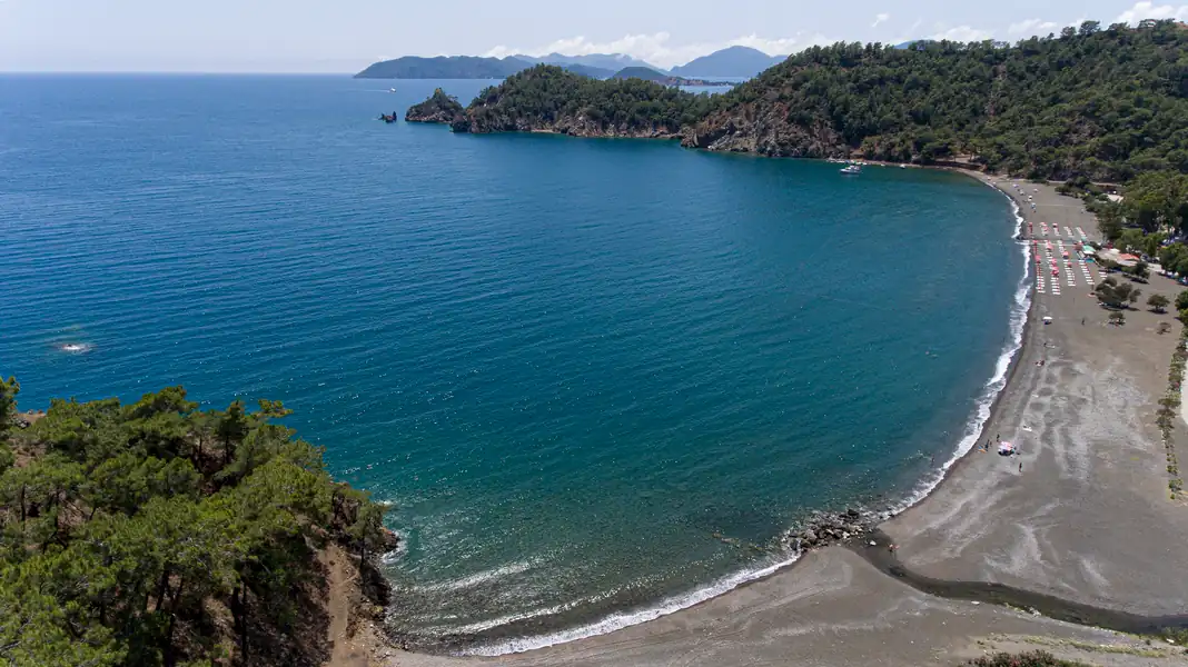 Göcek yakınlarındaki İnlice halk plajının sahil şeridinde yürüyüş yapan bir kişi. Çam ağaçlarının denize kadar uzandığı, sakin ve doğal bir ortam.